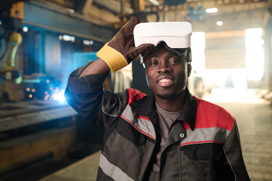 Happy young African American welder in workwear and protective gloves taking off vr headset and looking at camera against working machine
