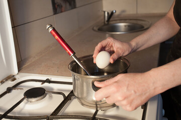 A girl boils egg in a metal pan on a white gas stove in the kitchen. Image for your creative decoration or design.
