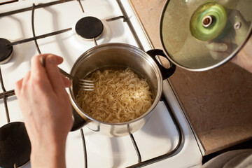 Hot instant pasta is cooked in a saucepan on the stove in the kitchen. The girl stirs the food in the bowl with a fork. Image for your creative decoration and design.
