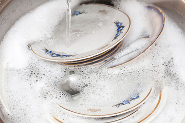 Composition about washing dishes. A set of large plates lie in the washbasin with foamy water. An...