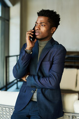 Young serious African American businessman in dark blue suit standing in front of camera and speaking to colleague on smartphone