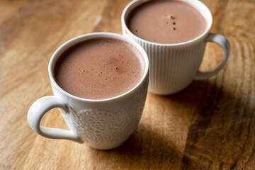 White mugs filled with rich, steaming cocoa on a rustic wooden table. The cozy and warm ambiance, coupled with the visual appeal of the white mugs and wooden texture, visual for coffee shops or cafes