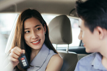 Happy Asian young woman showing remote car to her friend or boyfriend sitting in vehicle at automotive rental, customer satisfied feedback, test driving, dealership or purchasing concept, focus on key