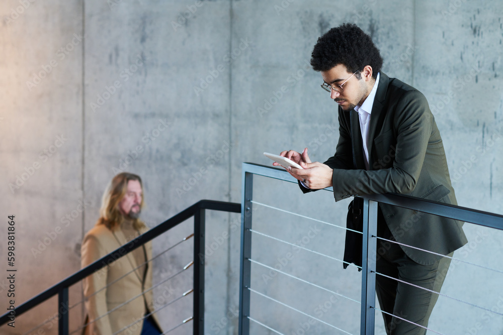Aufkleber young serious businessman looking through online data in tablet while standing against railing on st