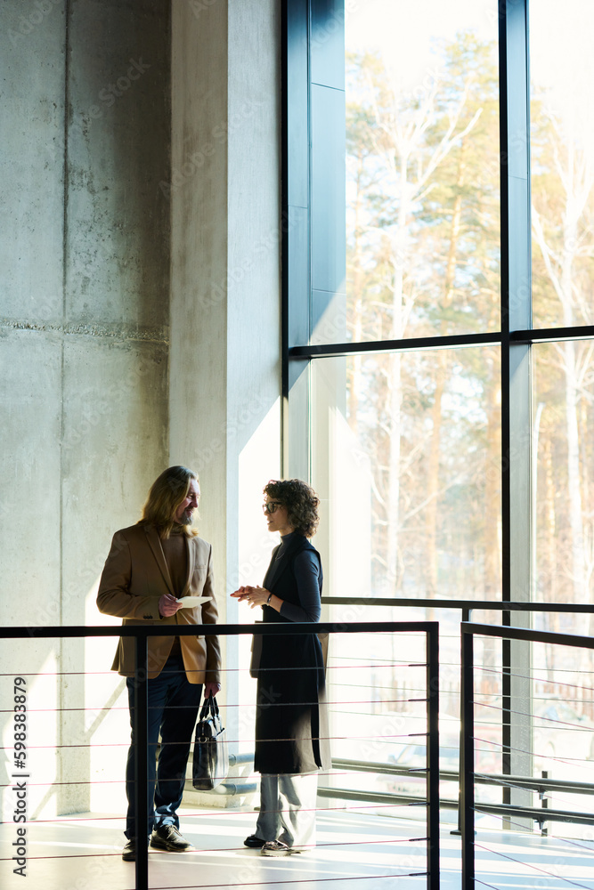 Poster Outlines of two mature colleagues discussing project points while standing in sunlit corridor of office center between window and railing