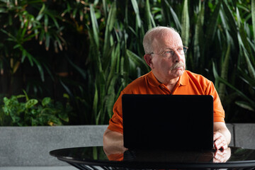 Grey haired senior man with mustache wearing eyeglasses and using laptop computer while thinking