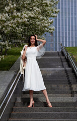 girl in a white dress stands on the stairs in a big city next to white flowering trees in spring