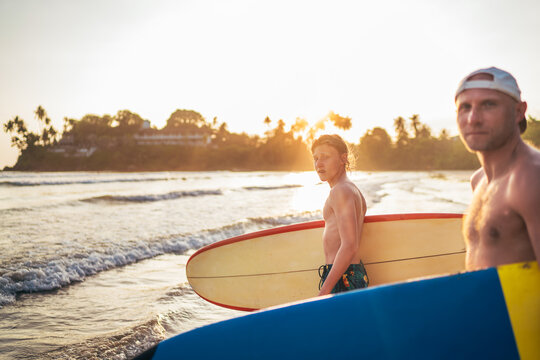 Teen boy with a surfboard goes to the sea surfing. He has a winter vacation and enjoying a beautiful sunset light with his father on Sri Lanka island. Family active vacation concept.