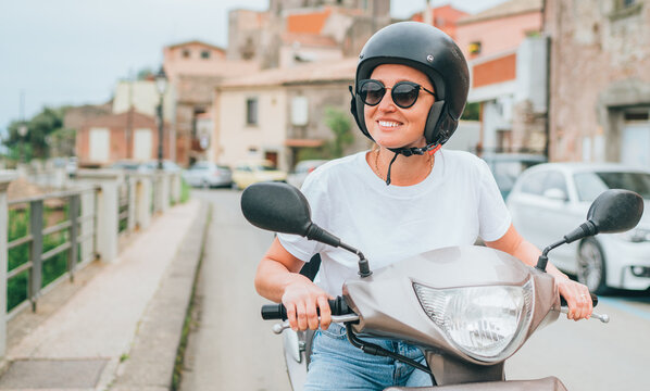 Cheerfully smiling woman in helmet and sunglasses fast riding the moto scooter on the Sicilian old town streets in the Forza d'Agro. Happy Italian vacation and transportation concept.