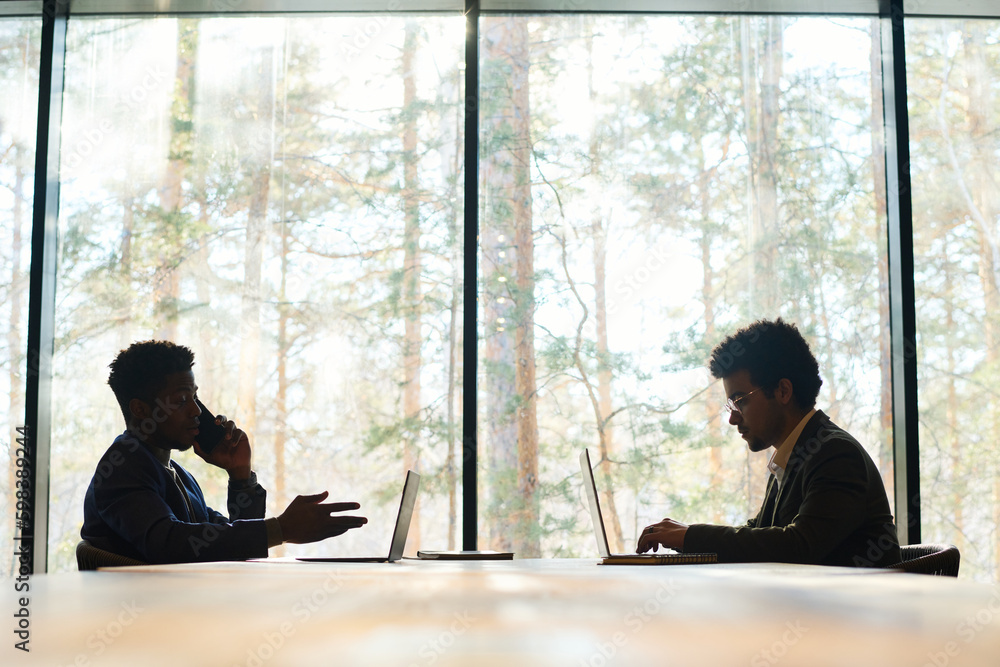 Poster Side view of two intercultural male coworkers using mobile gadgets while one of them typing on laptop and his colleague talking on smartphone