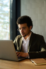 Fototapeta premium Young serious male broker or solopreneur typing on laptop keyboard and looking at screen while sitting by workplace and organizing work
