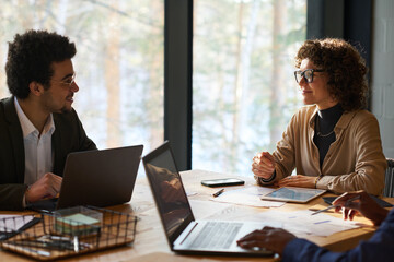 Two intercultural analysts with mobile gadgets consulting about financial points at meeting in boardroom while sitting in front of one another