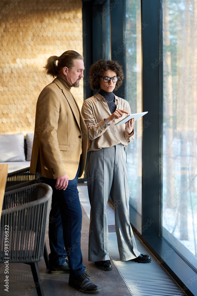 Poster Two confident analysts looking through online financial data in tablet held by brunette businesswoman and discussing it at meeting in office