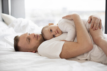 Obraz na płótnie Canvas Portrait of handsome bearded man holding little kid on belly. Kind father with adorable baby girl in white clothes lying on soft bedding and looking at camera. Concept of parental love and care.