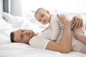 Portrait of handsome bearded man holding little kid on belly. Kind father with adorable baby girl in white clothes lying on soft bedding and looking at camera. Concept of parental love and care.