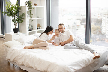 Young parents resting with pretty little daughter on comfy bed with panoramic windows on background. Loving mother and father embracing their toddler from both sides and smiling on camera.