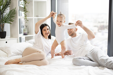 Loving mom and dad holding hands of active girl and helping baby to take first steps on large white...