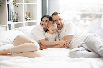 Positive father, mother and baby lying on wide bed of bright gorgeous apartment and looking at camera. Adoring parents hugging baby gently near big panoramic window. Sincere love concept.