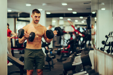 A young fit shirtless man is lifting dumbbells in a gym and working out his biceps in a sports center.