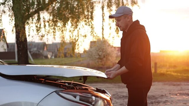Young man is opening the hood of a car outdoors in the evening on sunset. Driver checking or fixing the automobile engine on the road after a car breakdown. Vehicle service concept. Slow motion.