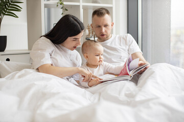 Fototapeta na wymiar Beautiful woman looking through wordless picture book together with baby girl and attentive male while resting under duvet in bedroom. Mindful parents telling stories to interested kid at weekend.