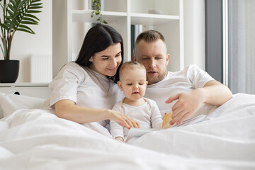 Fototapeta na wymiar Young married couple in casual outfit sitting in cozy bed with little daughter and reading interactive baby book on Sunday morning. Loving parents making story come alive for curious daughter.