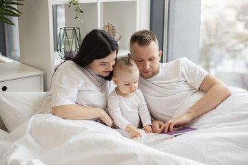 Obraz premium Young married couple in casual outfit sitting in cozy bed with little daughter and reading interactive baby book on Sunday morning. Loving parents making story come alive for curious daughter.