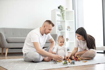 Caring mother and father in casual clothes resting on floor while helping little baby daughter with...