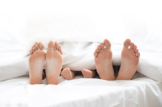 Close up view of three pairs of bare feet being hidden under white soft blanket on comfortable bed indoors. Happy mother, father and infant baby relaxing together during sunny morning at home.