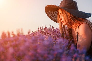 Woman lavender field. Happy carefree woman in black dress and hat with large brim smelling a blooming lavender on sunset. Perfect for inspirational and warm concepts in travel and wanderlust. Close up - obrazy, fototapety, plakaty