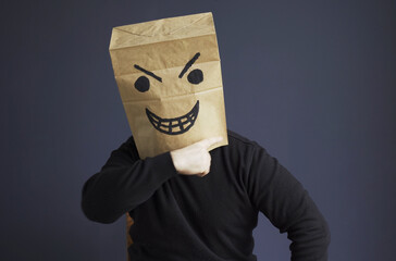 A man in a black turtleneck with a paper bag with an angry emoticon on his head shows a threatening gesture.