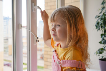 safety at home for small children. a lock on the window protects children from opening the window. The girl 4 years old tries to open the window standing on the windowsill on a high floor.