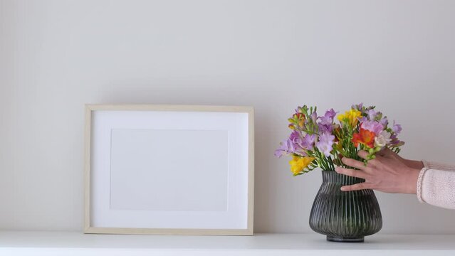 Female hands puts flowers in a vase. Wooden frame mockup in white room interior, empty mock-up with copy space. Light frame with a vase in the interior for design presentation. Generative AI