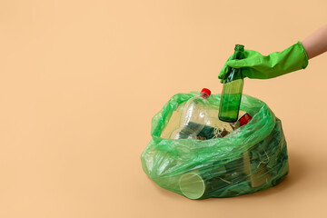 Fototapeta na wymiar Female hand in rubber glove putting glass bottle into garbage bag on beige background