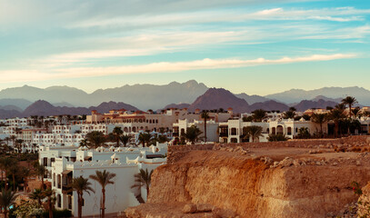 Imposing desert mountains provide a stunning backdrop to a cluster of white buildings, nestled amidst flourishing palm trees, capturing the essence of a tranquil desert oasis