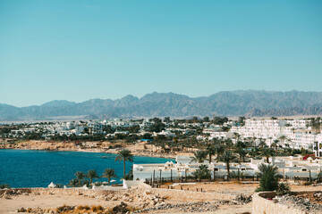 An enchanting view unfolds as azure waters meet endless desert sands, adorned by pristine white buildings and swaying palm trees, a picturesque oasis.
