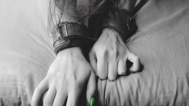 Girl's hands are shackled with red leather handcuffs for role-playing close-up. Gets pleasure, squeezing the sheets with his hands. Black and white
