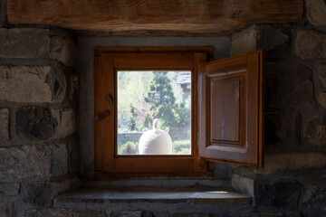 a white botijo seen from the inside of a window of a stone village house with wooden shutters, typical spanish clay botijo to keep the water cool in summer, rustic atmosphere