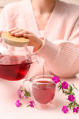 Woman pouring hot tea from teapot into cup with delicate flowers on pink table