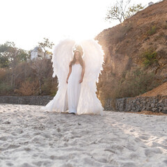 Beautiful angel with big white wings. Young tender woman in white silk dress walking at seashore