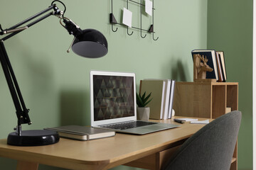 Modern laptop, books, lamp and stationery on wooden desk near green wall. Home office