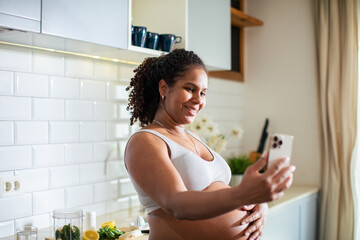 Fototapeta na wymiar Young pregnant latina woman using a smart phone and having a video call while in the kitchen of a house