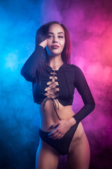 Young beatiful woman in black underwear posing against a background of blue and pink smoke from a vape on a black isolated background