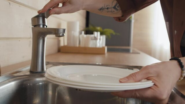 Unrecognizable woman bringing plates to wash, turning on faucet and getting no water, water cut-off concept