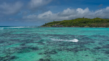 The waves are foaming in the boundless turquoise ocean. Corals are visible through the clear water. The villas of the hotel are visible on the shore of the green island. Blue sky, clouds.Seychelles.