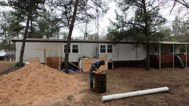 Man cutting round hole in the rusted metal barrel next to pile of sand dug from home made water well project.