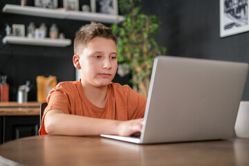 Teen boy study or play game on laptop home interior background. Guy doing homework and typing on laptop