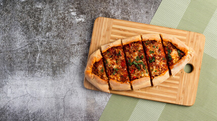 Sliced Pide (turkish pizza or pita dish) with ground beef, cheese, tomatoes and greens on a rectangular cutting wooden board on a dark grey background. Top view, flat lay