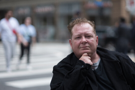 With pedestrians in the background,, a disabled transman in a powerchair pauses in a crosswalk to express the different ways that people interact with public space and accessible design.