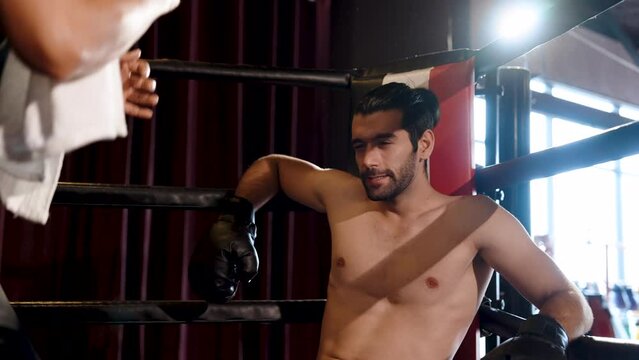 Strong Caucasian sportsman boxer is sitting in corner of a boxing ring with his mentor coaching him on how to fight deliberately in order to defeat his opponent in a single salute. in boxing stadium